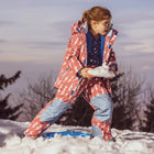 A young girl in a Blizzard Kids Ski Jacket Pink, holding snow outdoors. She's wearing sunglasses and a patterned ski suit.